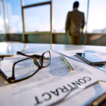 Business contract, pen, eyeglasses, market analysis and cellphone on desk and businessman standing on background