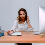 Young concentrated woman in headphones and microphone talking with client while sitting at the office desk over white background