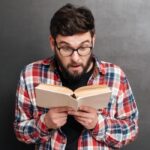 Surprised man dressed in shirt in a cage and wearing glasses standing over chalkboard while reading a book