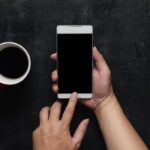 Hand using phone black screen with cup of coffee. Wooden black office desk table on top view .