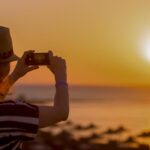 Young woman in straw hat and cute summer dress standing at the balcony with picturesque sea scenery, taking picture of sunset or sunrise on smartphone, back view